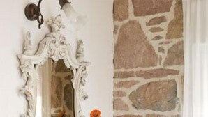 A room with a stone wall, a white ornate mirror, a small white vase with a flower, a wooden table with a coffee pot, and a newspaper. - Cunda Lena Otel (Ayvalik)