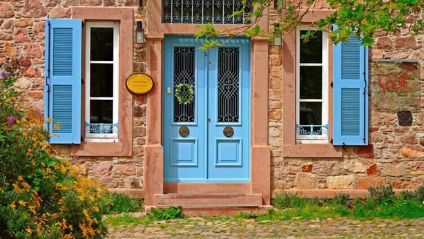A stone building with a blue door, two windows with blue shutters, and a small garden area with flowers. - Cunda Lena Otel (Ayvalik)