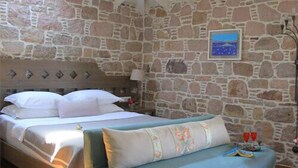 A bedroom with a stone wall, a bed with white linens, a blue sofa, and a nightstand with a lamp. - Cunda Lena Otel (Ayvalik)