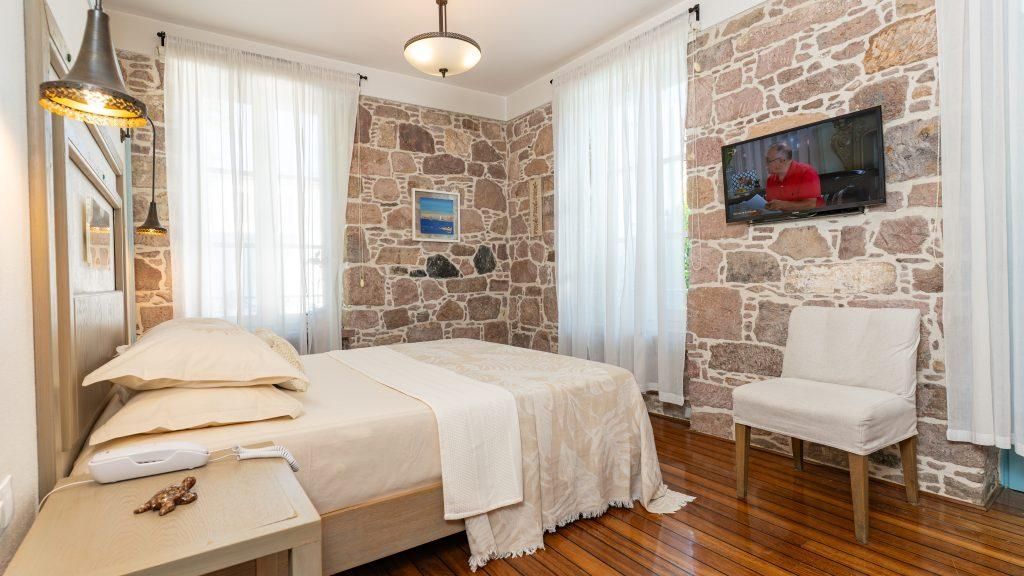 Een slaapkamer met een stenen muur, een bed met een witte spreideken, een stoel, een aan de muur bevestigde televisie en een nachtkastje met een lamp en een afstandsbediening.
