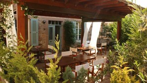 An outdoor patio with wooden tables and chairs, surrounded by potted plants and a stone wall. - Cunda Lena Otel (Ayvalik)