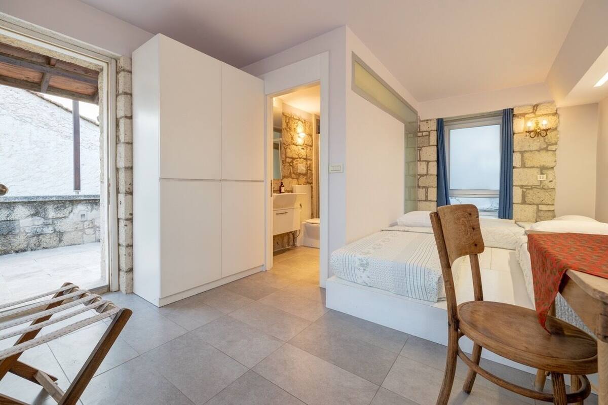 A compact room with a bed, a wooden chair, a table, and a view of a stone wall and window.
