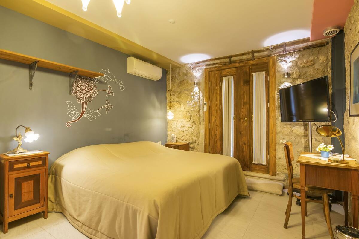 A bedroom with a bed, bedside tables, a desk, a chair, a TV, and a stone wall.