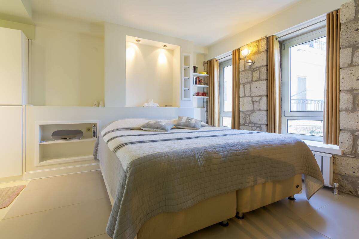 A bedroom with a bed, built-in shelves, a window with curtains, and a stone wall.