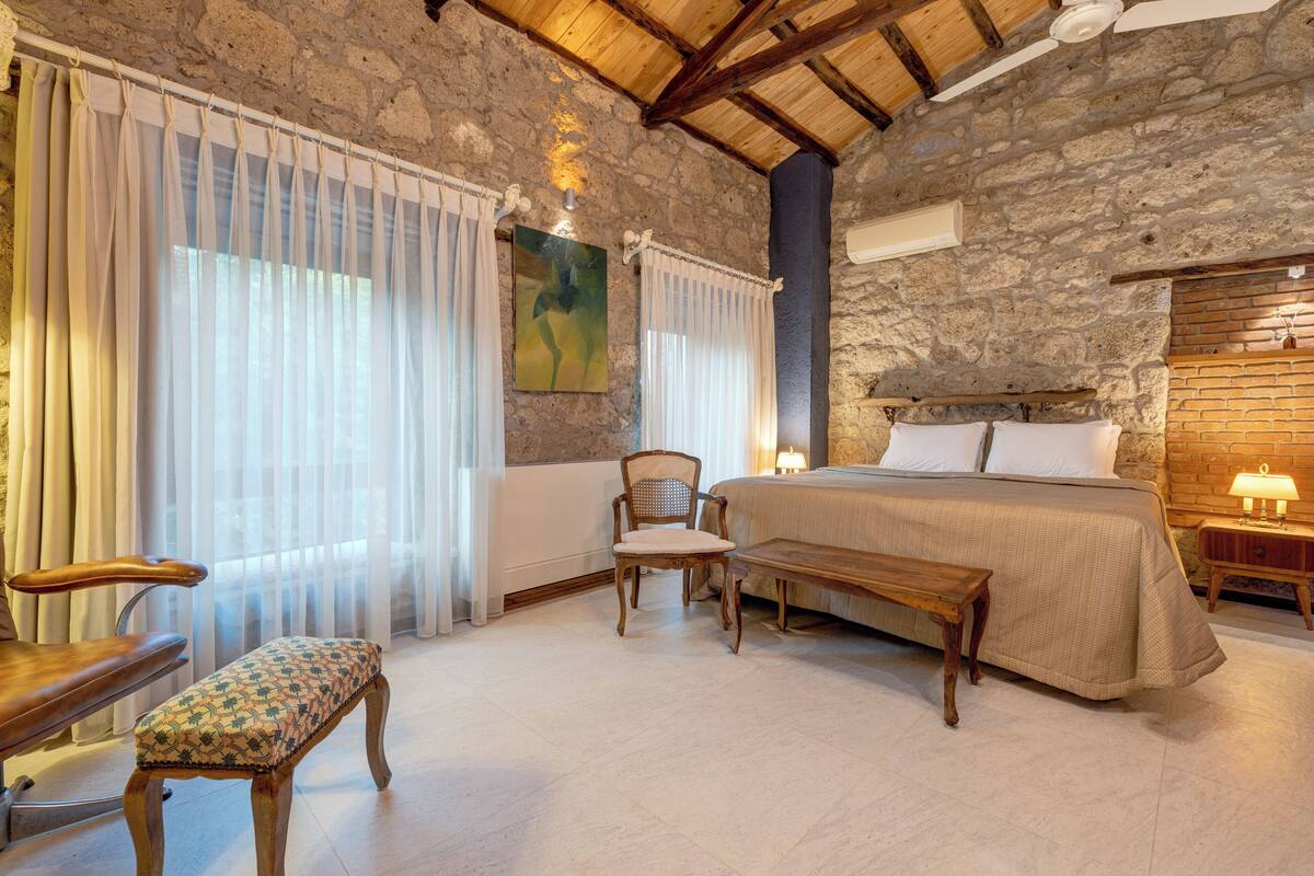 A bedroom with a stone wall, a wooden bed, a bench, a chair, and a bedside table with a lamp.