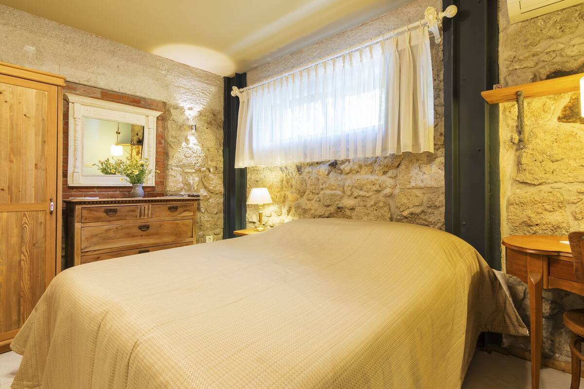A bedroom with a large bed, wooden furniture, a stone wall, and a window with curtains.