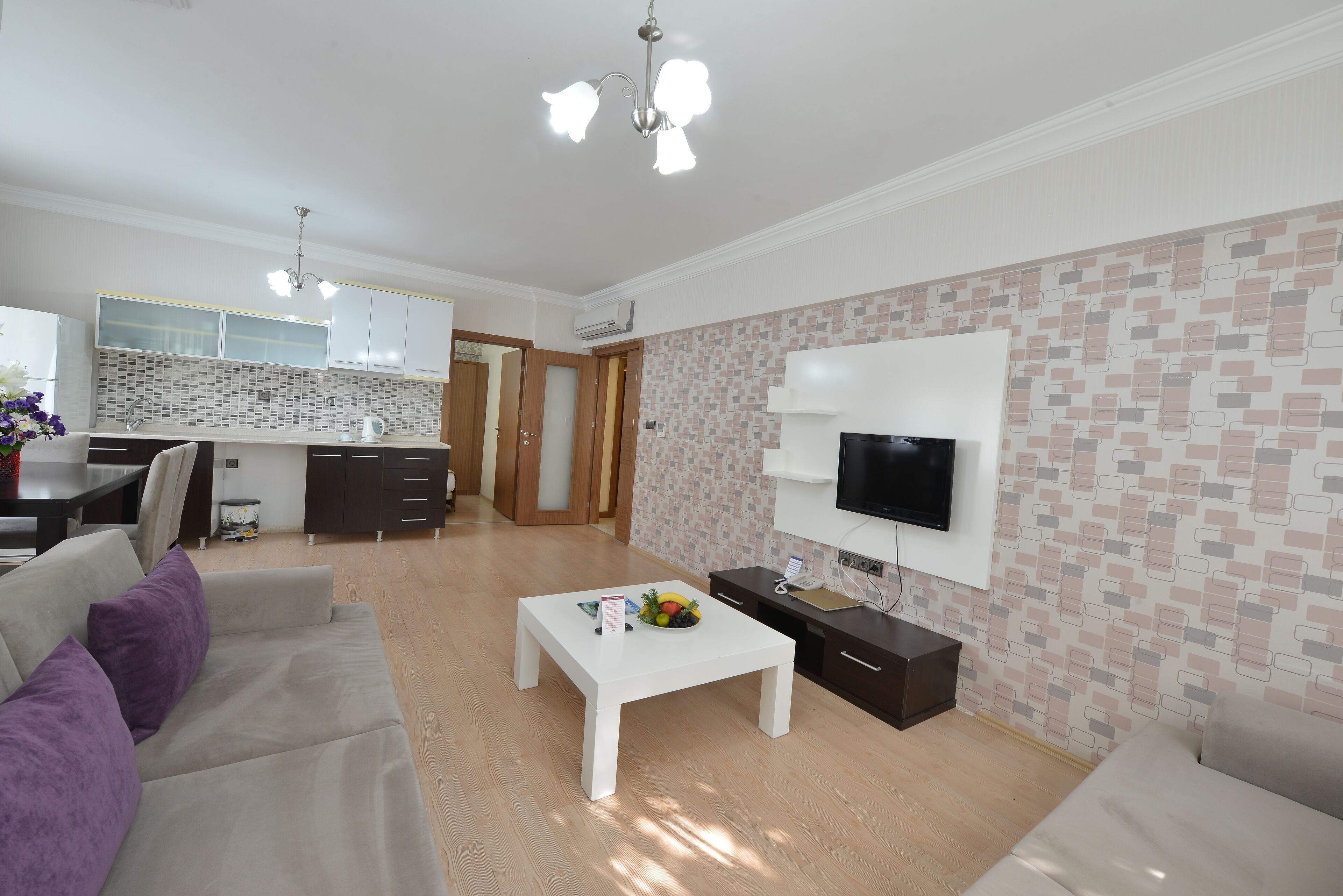 Una sala moderna con un sofá blanco, una mesa de centro con fruta, un televisor de pantalla plana y una cocina al fondo.