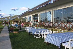 An outdoor dining area with tables and chairs, a pathway, and a building with large windows. - Riva's Club (Çekmeköy)