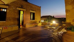 A courtyard with a tiled floor, a covered seating area with chairs, and a building with a door and windows. - Villa Laina (Platanias)
