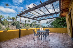 Un patio coperto all'aperto con tavolo e sedie, un muro giallo e vista su palme e colline.