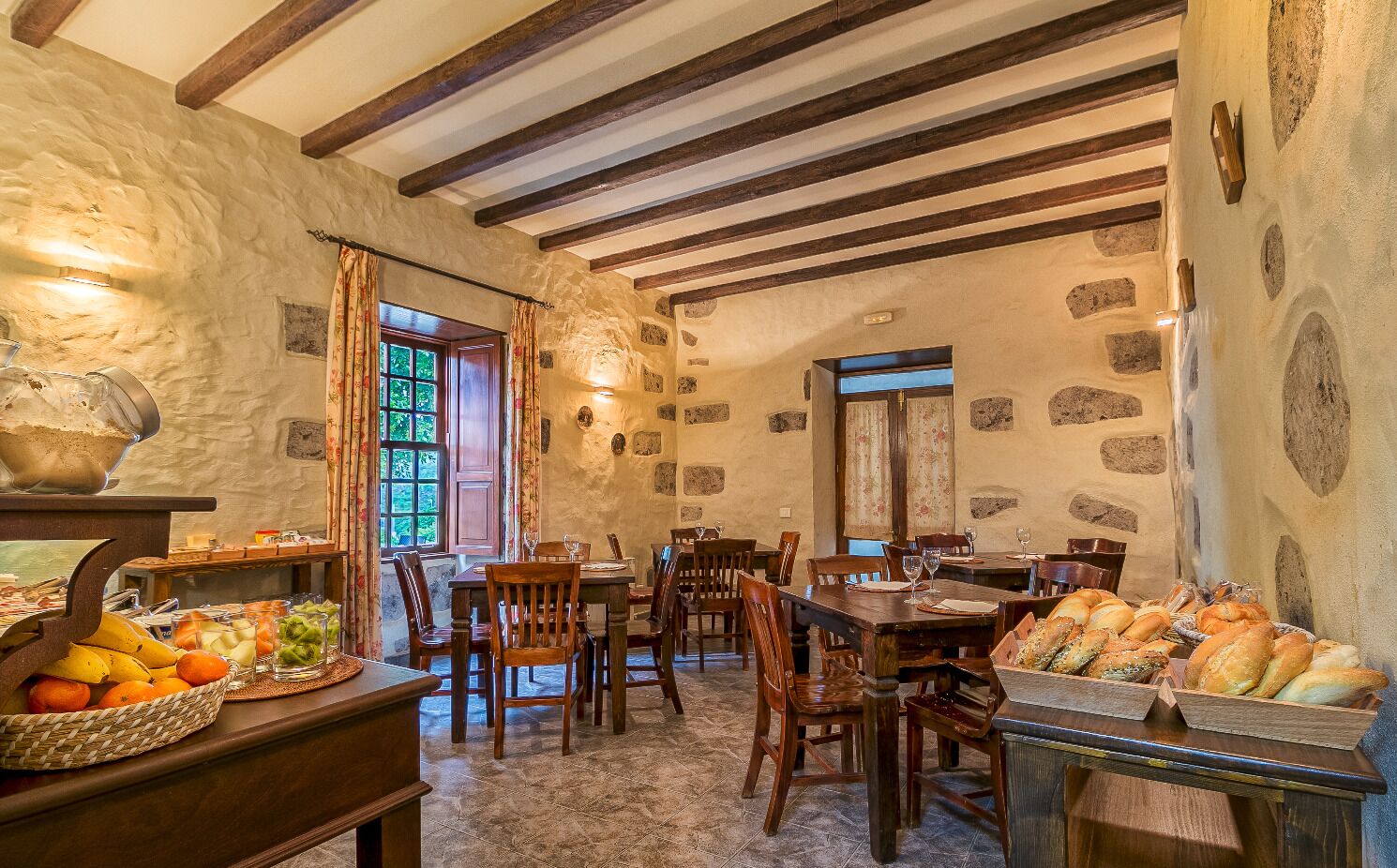 A rustic dining area with stone walls, wooden beams, and a variety of bread and fruits on the tables.