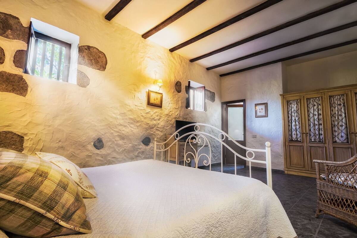 A bedroom with a bed, a window, a wall with stone-like texture, a wooden wardrobe, and a wicker chair.