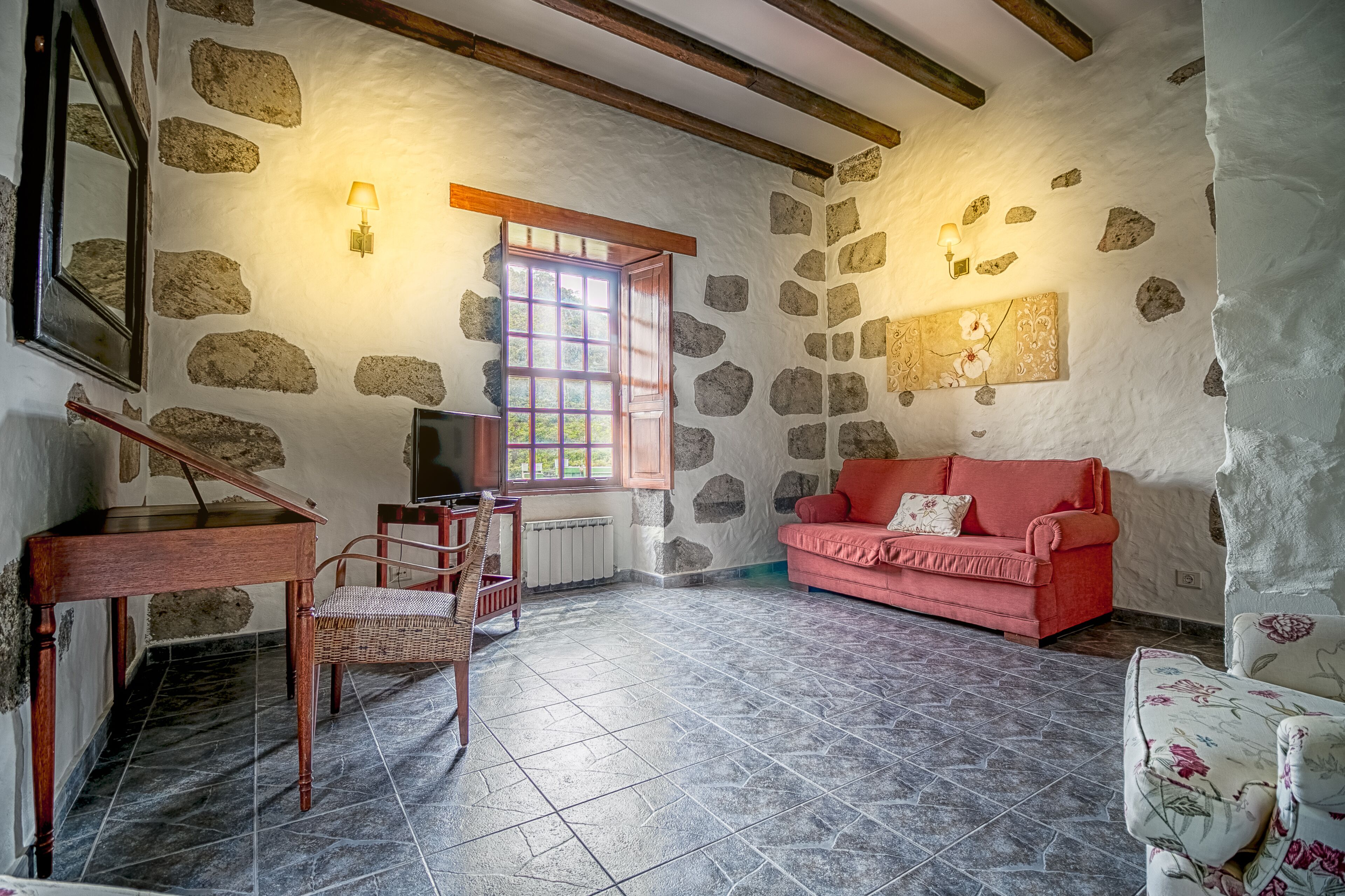 A rustic room with stone walls, a wooden ceiling, a red sofa, a wicker chair, and a piano.
