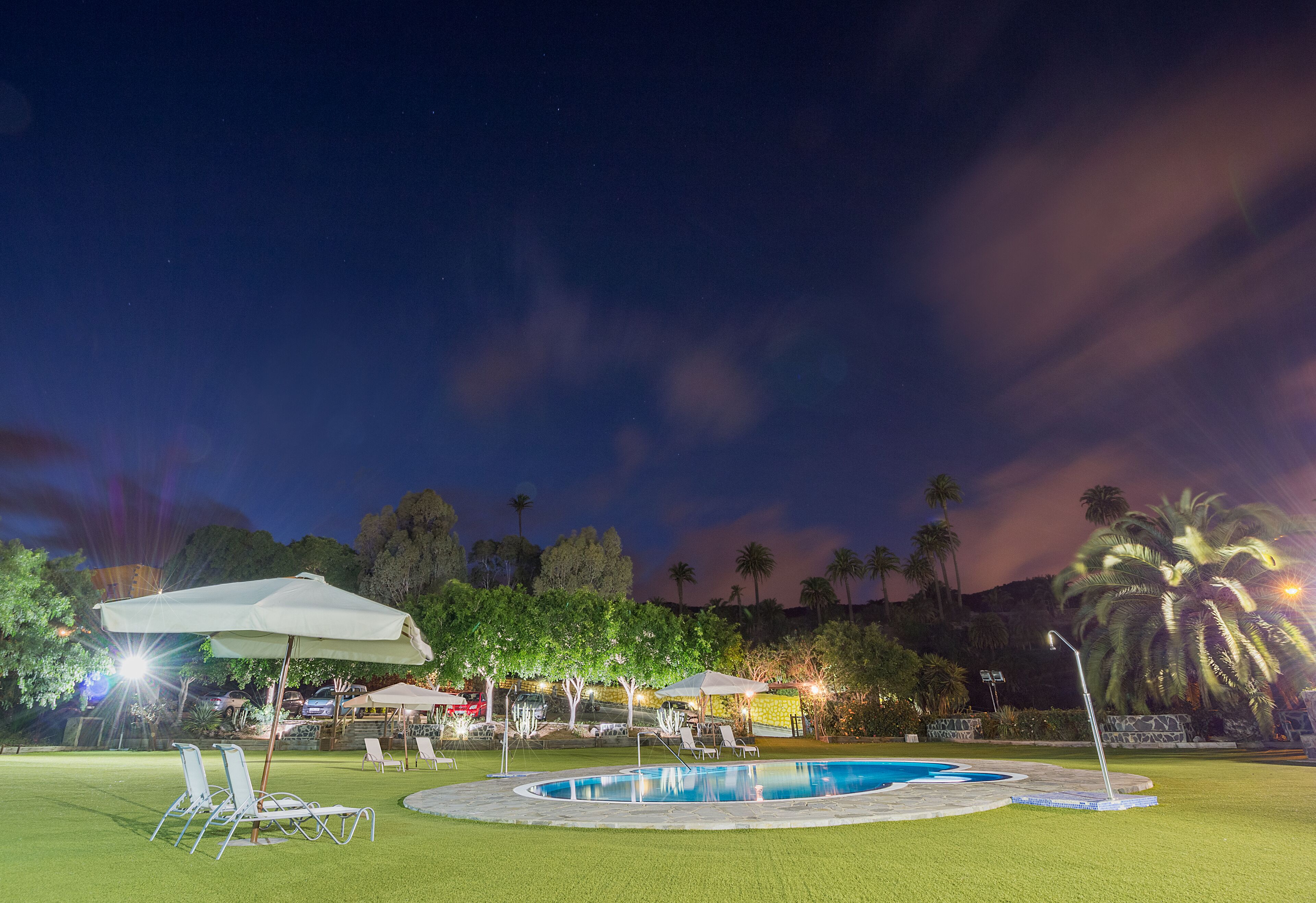 A nighttime pool area with lounge chairs and umbrellas, surrounded by palm trees and a well-lit environment.