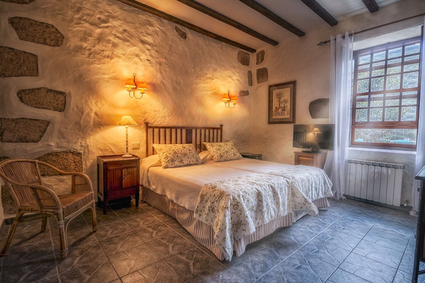 Um quarto aconchegante com parede de pedra, cama de madeira com roupa de cama floral, uma cadeira de vime e uma janela com cortinas.