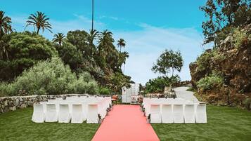 Un pasillo alfombrado rojo que conduce a un escenario con sillas blancas a ambos lados, rodeado de follaje y palmeras.