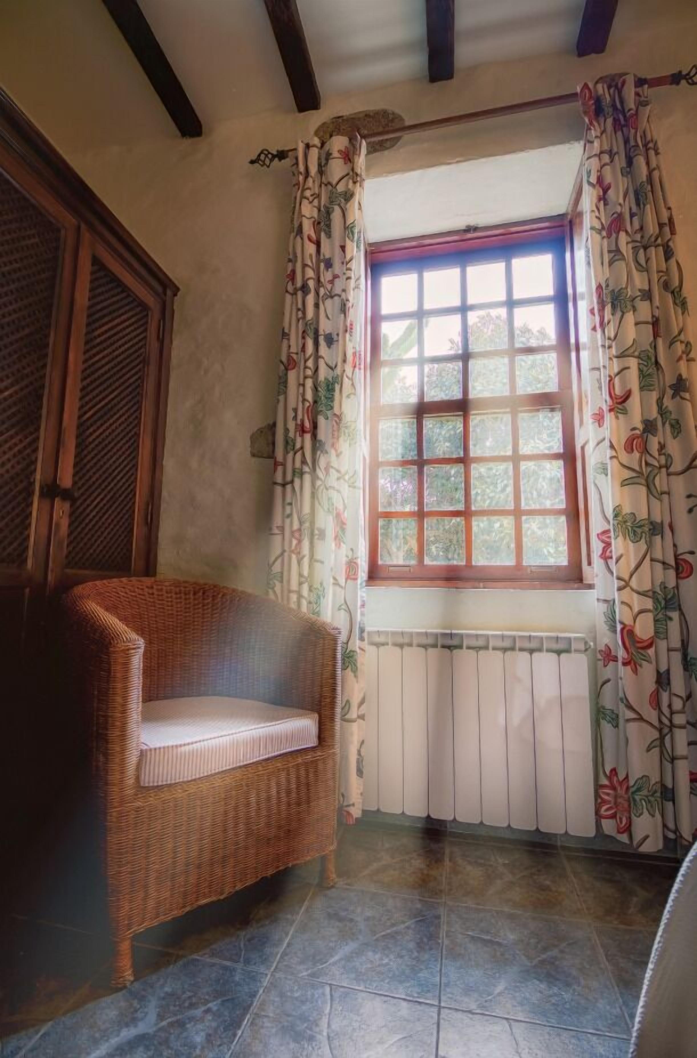 Una habitación de esquina con una silla de mimbre, una ventana con cortinas florales y un radiador.