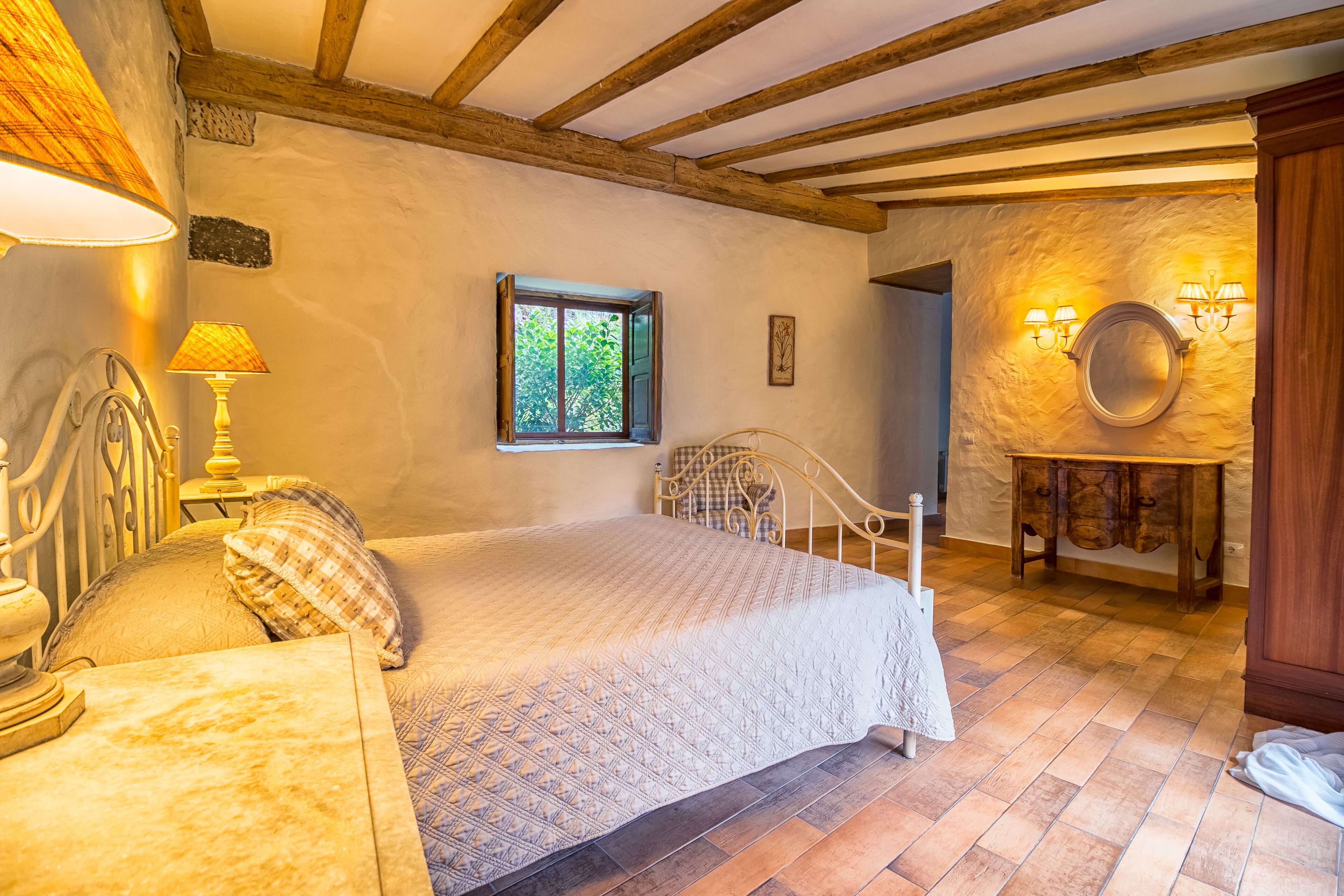 Un dormitorio con cama, mesita de noche, lámpara, ventana y espejo adosado a la pared.