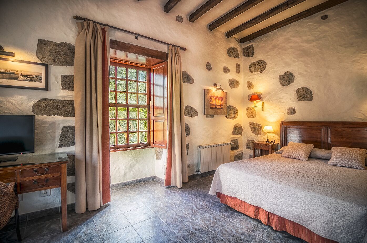 Un dormitorio acogedor con pared de piedra, una cama de madera, una mesita de noche, un televisor y una ventana con cortinas.