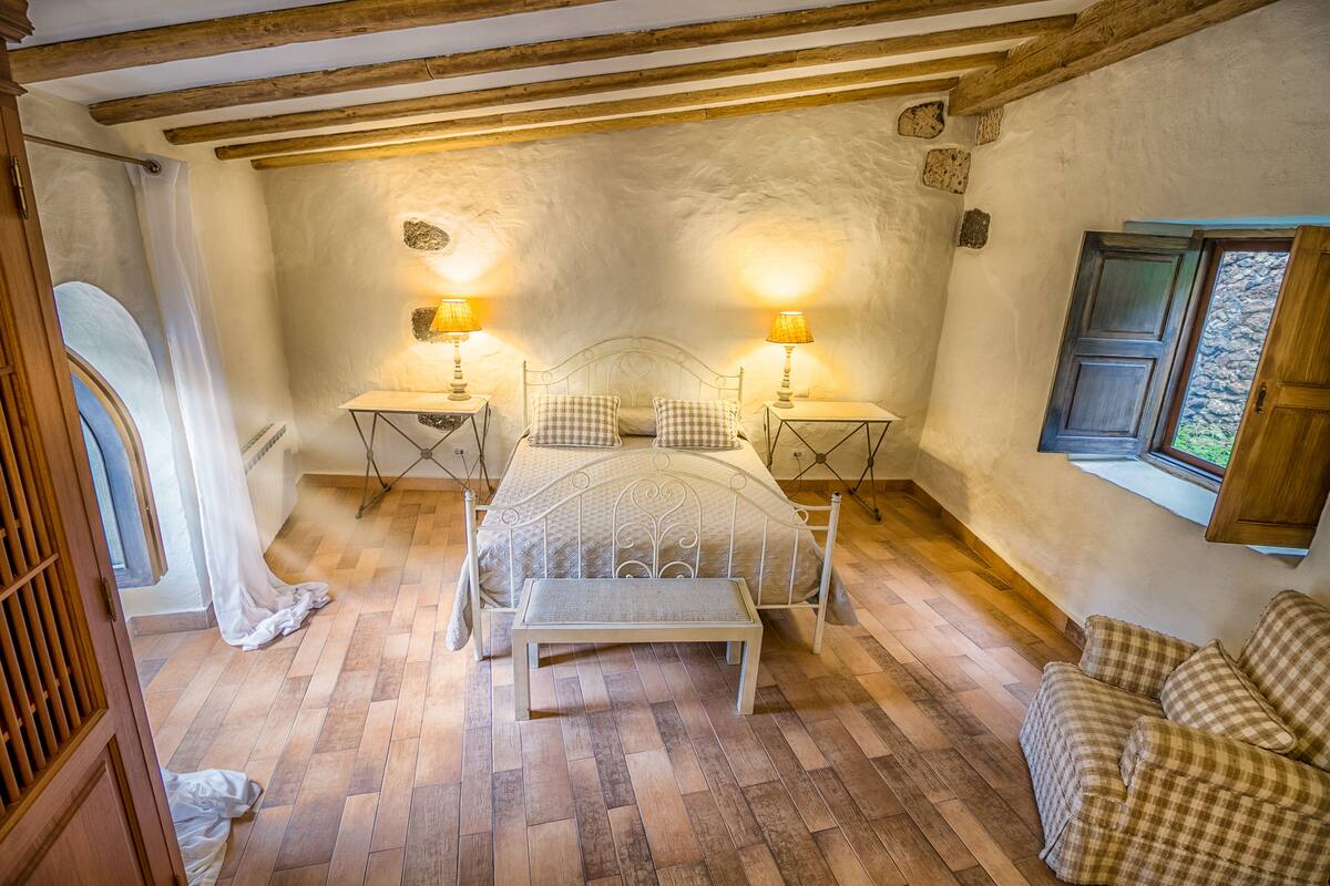 A bedroom with a bed, two bedside tables with lamps, a bench, a wooden floor, and a window with curtains.