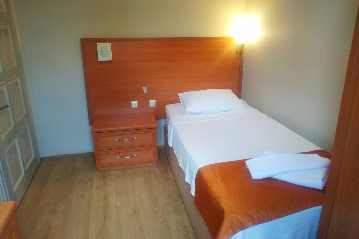 A small, neatly arranged bedroom with a wooden headboard, a single bed with white bedding, a nightstand with drawers, and a wall-mounted light.