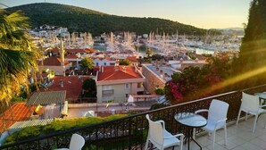 Un balcón con sillas y una mesa blancos, con vistas a una marina llena de barcos y una colina al fondo.