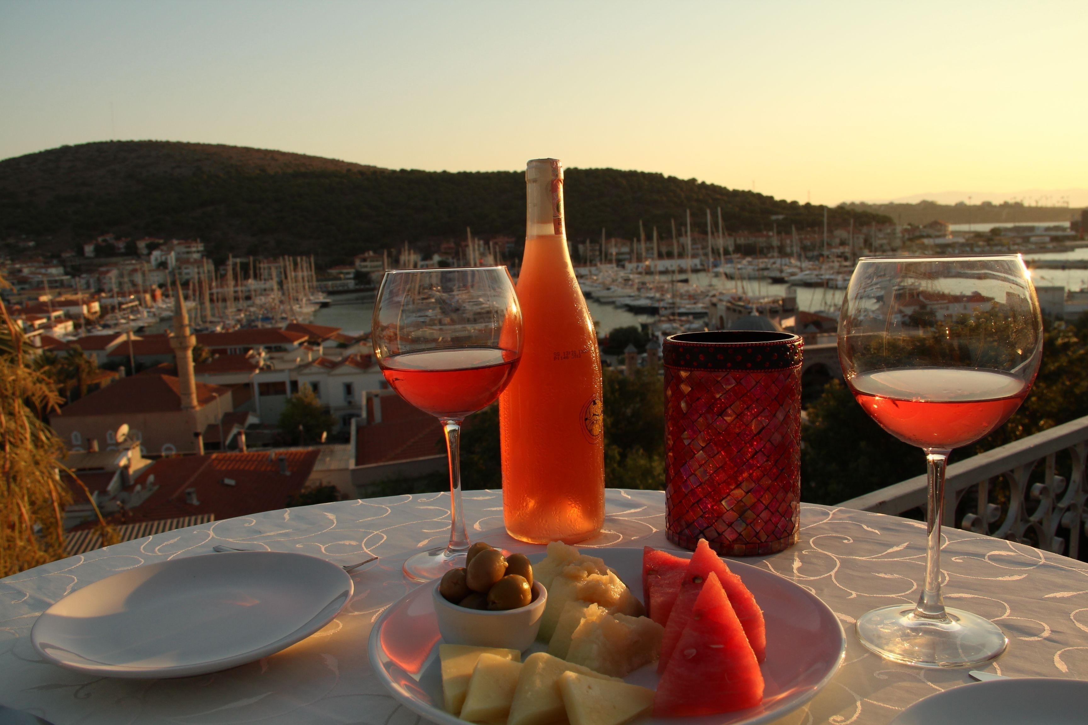 A table with a bottle of water, two glasses of red wine, a bowl of olives, and a plate with fruit on a balcony overlooking a marina and a hilly landscape.