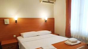 Habitación de hotel con cabecera de madera, dos lámparas de noche, aire acondicionado, cama con ropa blanca, mesita de noche con cajones y ventana con cortinas.