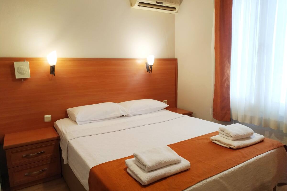 A hotel room with a wooden headboard, two bedside lamps, an air conditioner, a bed with white linens, a nightstand with drawers, and a window with curtains.