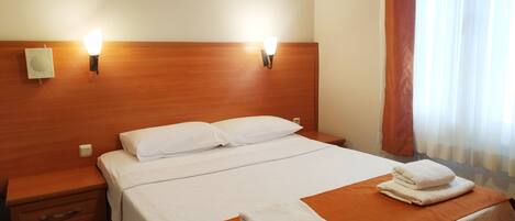 A hotel room with a wooden headboard, two bedside lamps, an air conditioner, a bed with white linens, a nightstand with drawers, and a window with curtains.