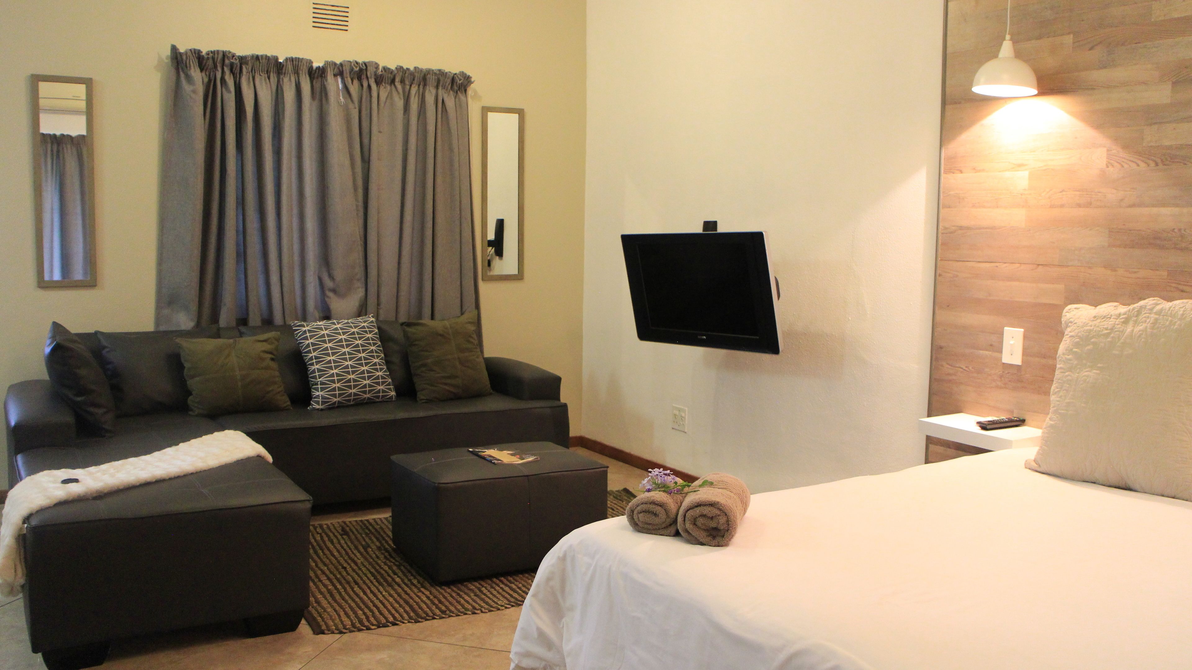A hotel room with a sofa, a TV mounted on the wall, a bed with a white bedspread, and a small table with towels.