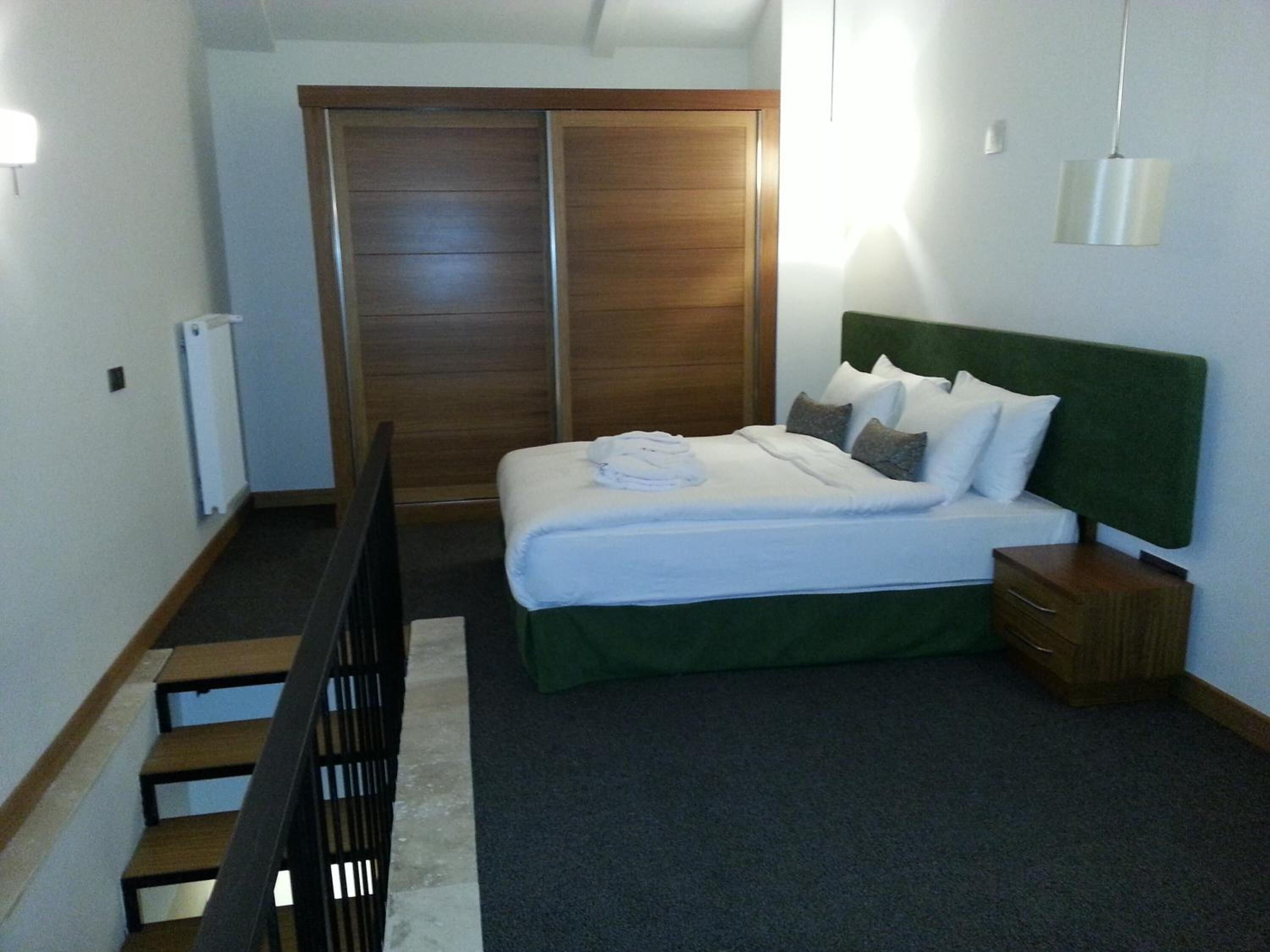 A bedroom with a bed, a nightstand, a wooden wardrobe, and a staircase.