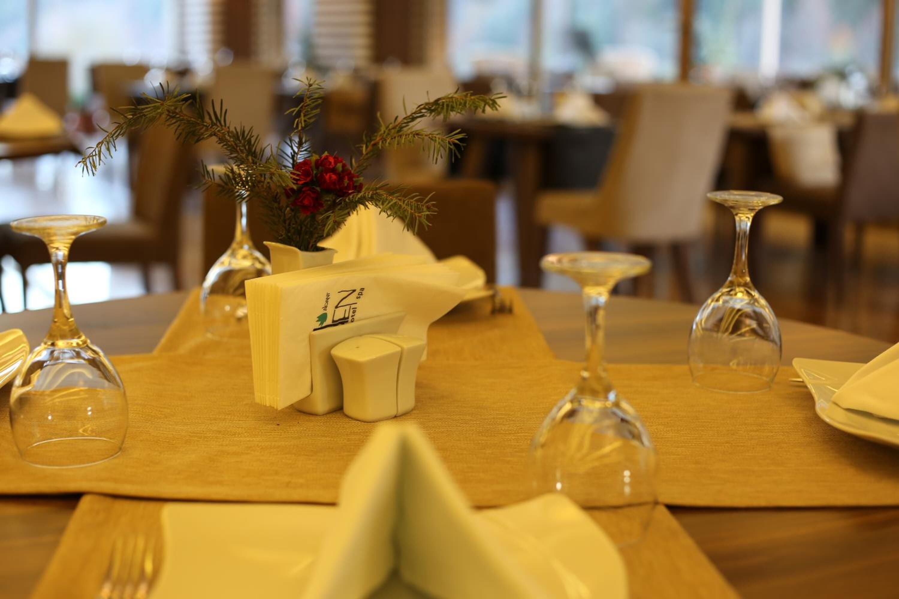 Una mesa de comedor dispuesta con un mantel amarillo, copas de vino y un centro de mesa con follaje y flores rojas.