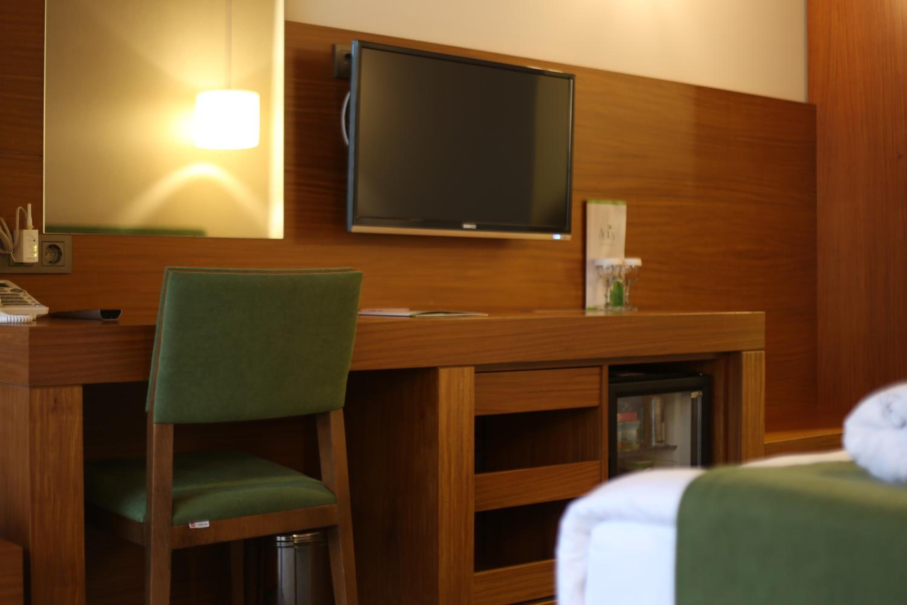 A hotel room with a wooden desk, a green chair, a television mounted on the wall, and a bed with a white blanket.