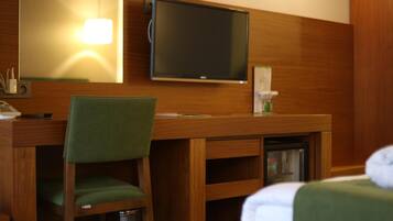 Habitación de hotel con un escritorio de madera, una silla verde, un televisor montado en la pared y una cama con una manta blanca.