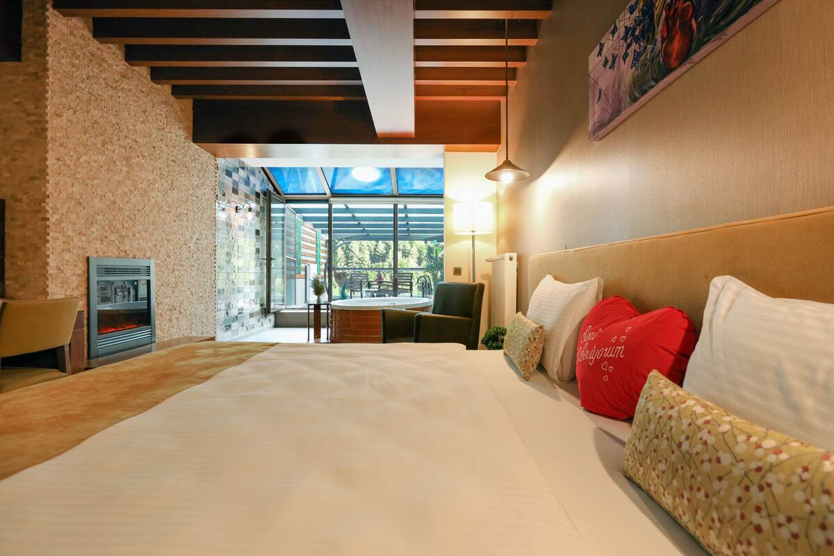 A hotel room with a large bed, a fireplace, a dining area with a view, and a modern ceiling design.