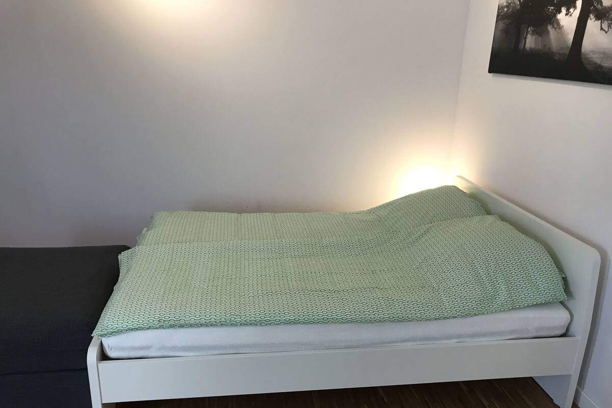 A single bed with a patterned green blanket, a white headboard, and a wall-mounted picture.