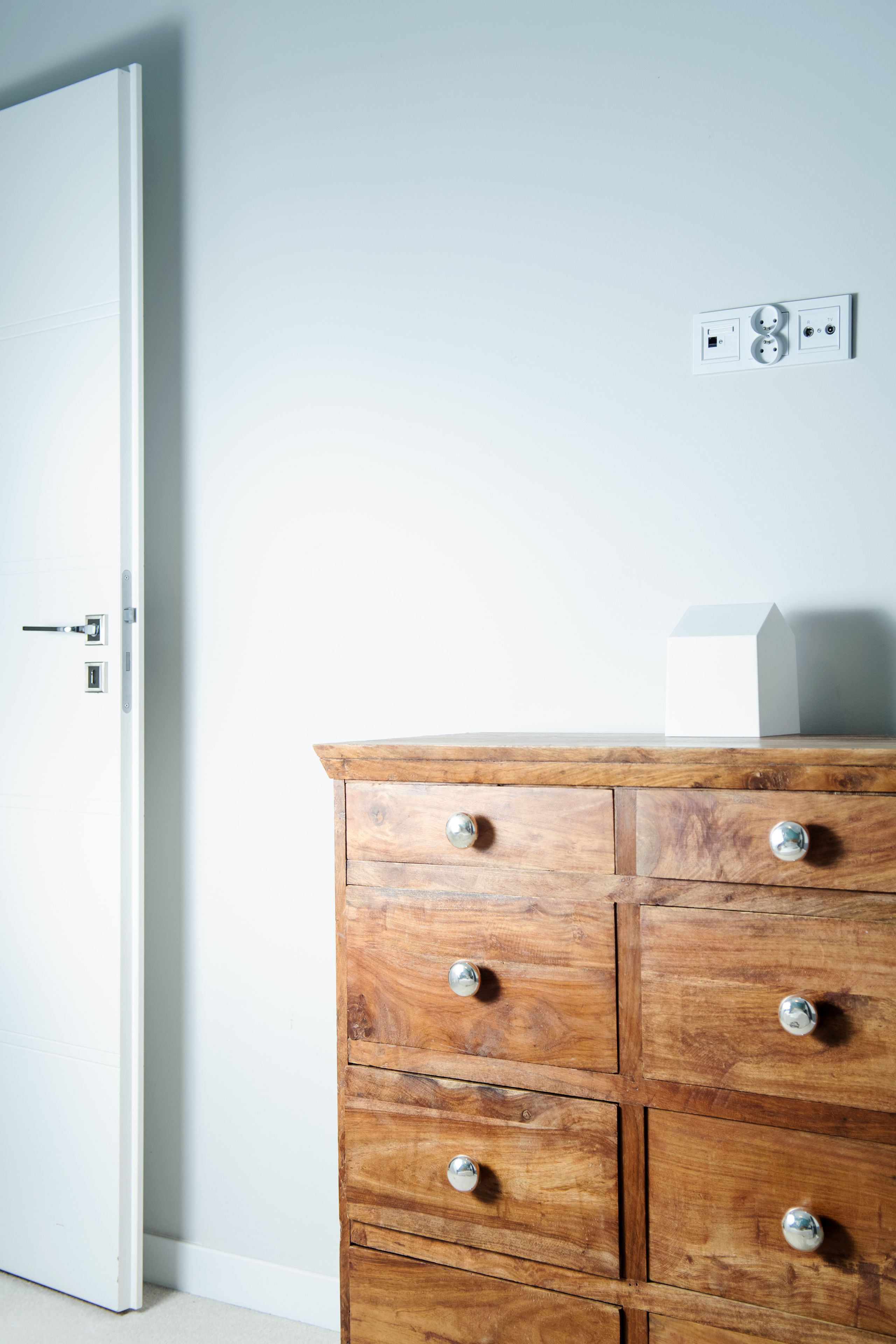 A wooden dresser with drawers and a small white object on top, a white door with a handle, and a wall socket with two plugs.