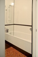 A bathroom with a glass-enclosed shower, a white bathtub, and a wooden mat on the floor.
