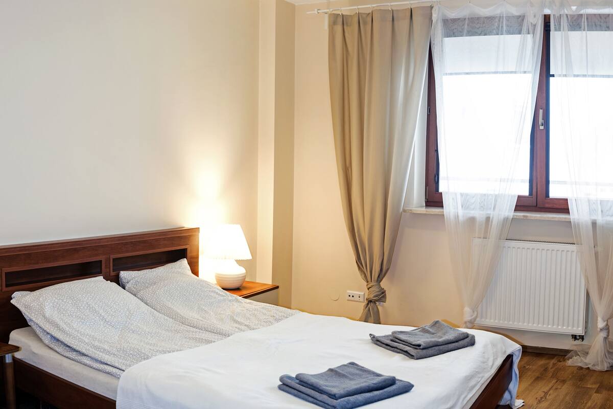 A neatly made bed with a white and patterned blanket, a bedside lamp, and a window with sheer curtains.
