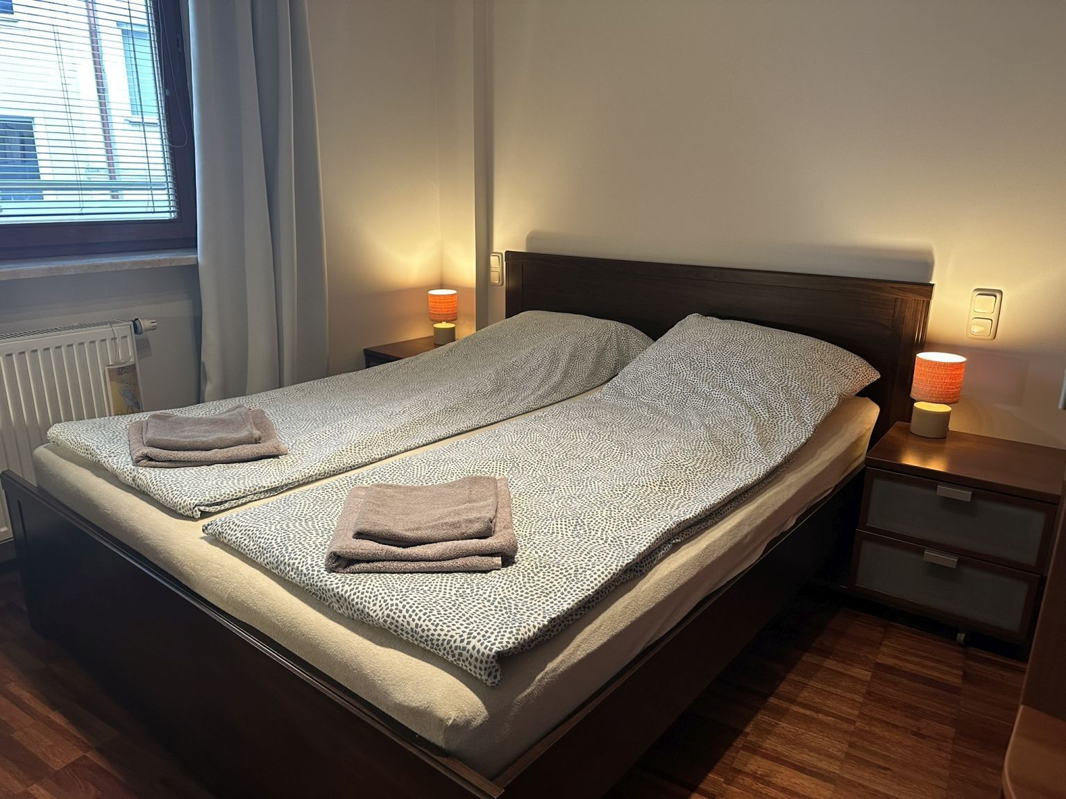Un lit double avec une couvre-lit à motifs, deux serviettes, des lampes de chevet, une table de chevet et une fenêtre avec des stores.