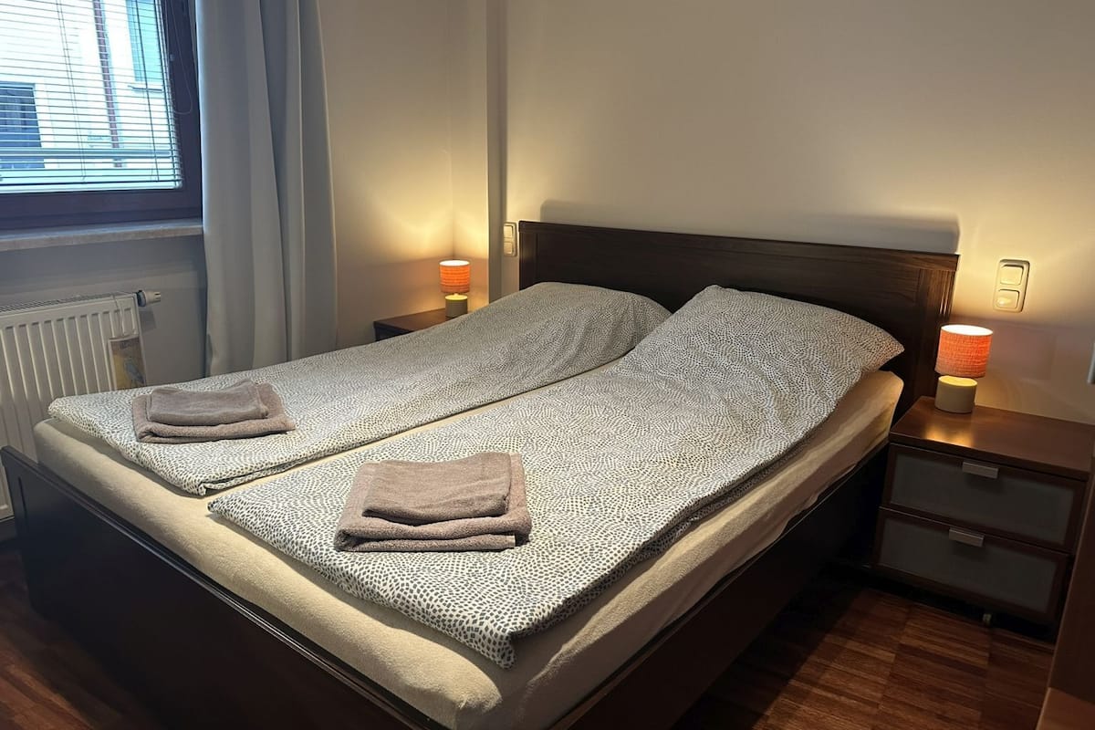 A double bed with a patterned bedspread, two towels, bedside lamps, a nightstand, and a window with blinds.