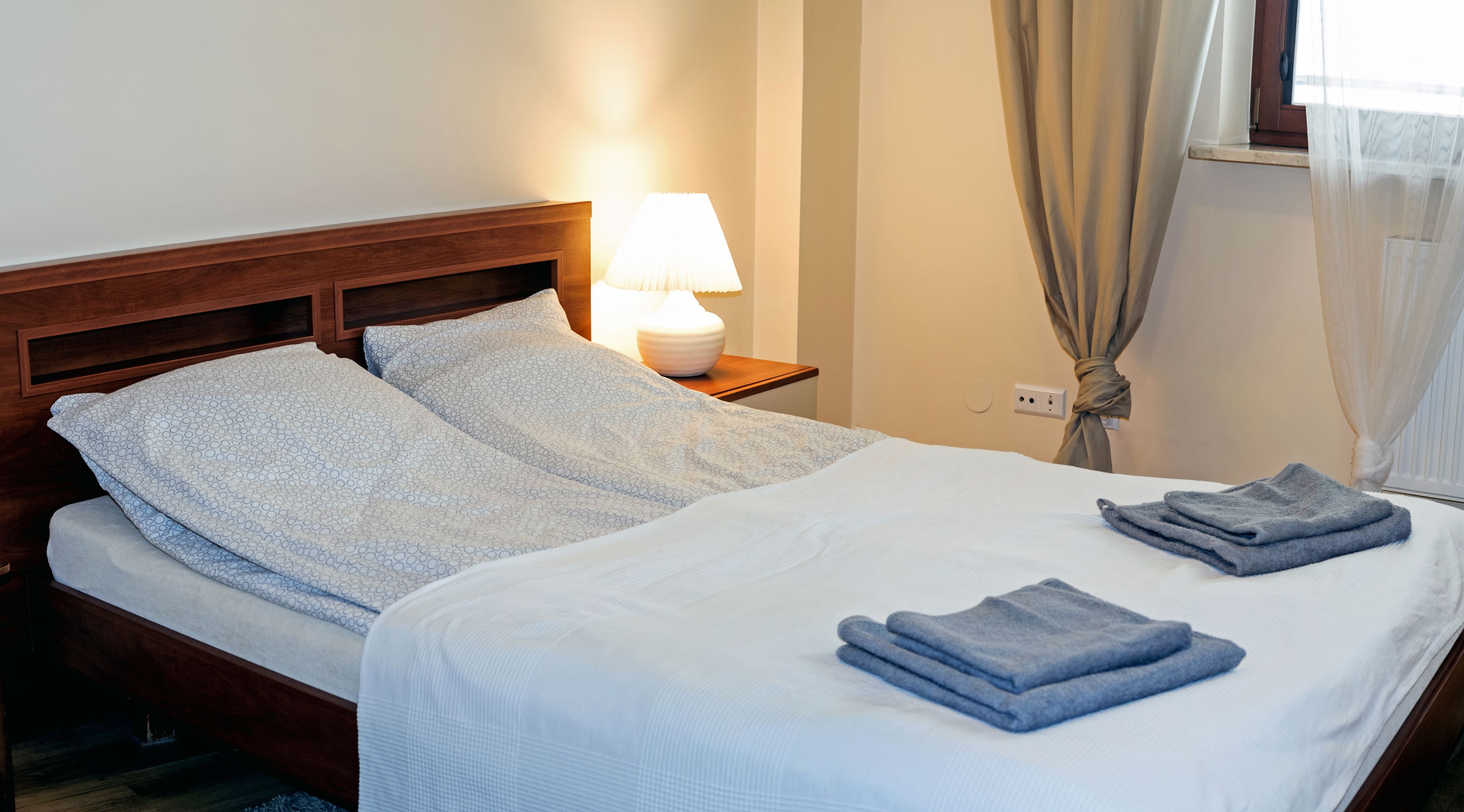 A neatly made bed with a white and blue patterned bedspread, a wooden headboard, a bedside lamp, and a folded towel on the bed.