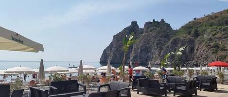 An outdoor seating area with tables and chairs, umbrellas, and a view of the sea and a rocky cliff.