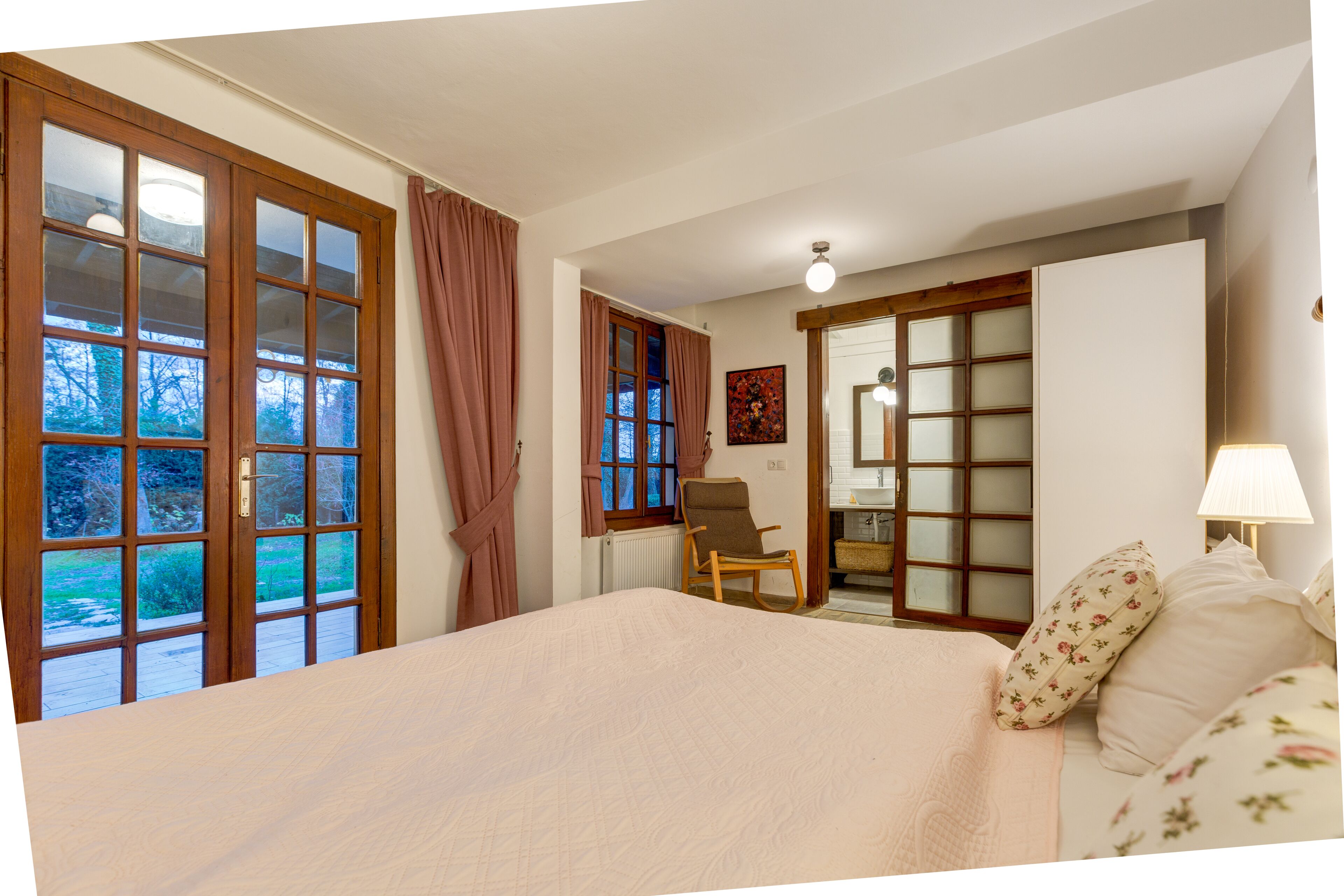 A bedroom with a large bed, a wooden rocking chair, a mirror, and a view of the outdoors.