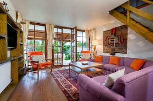 A living room with a purple sofa, orange armchairs, a wooden coffee table, and a large window with curtains.