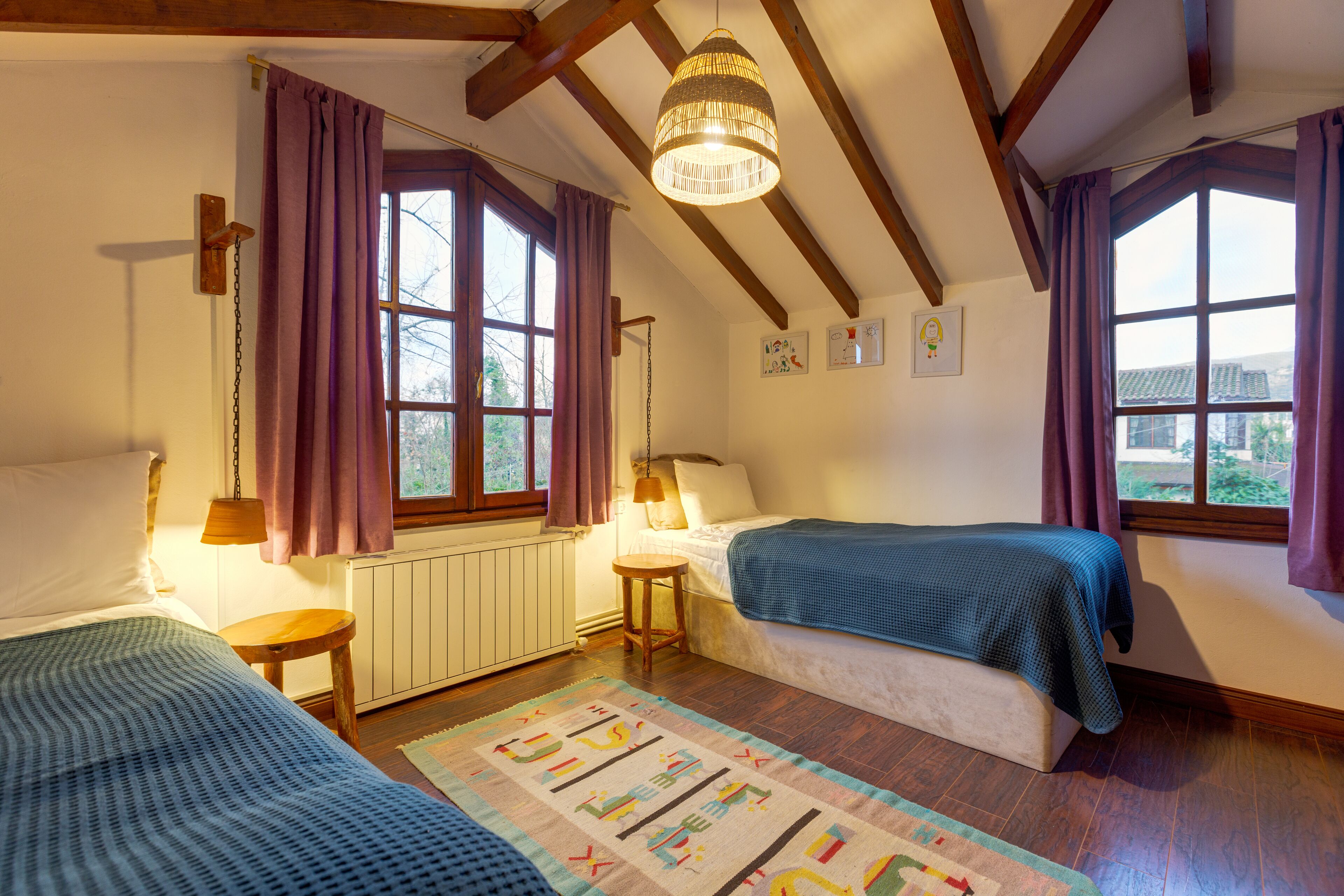 Een knus slaapkamer met twee eenpersoonsbedden, houten balken, een groot raam met paarse gordijnen, een hanglamp en een radiator.
