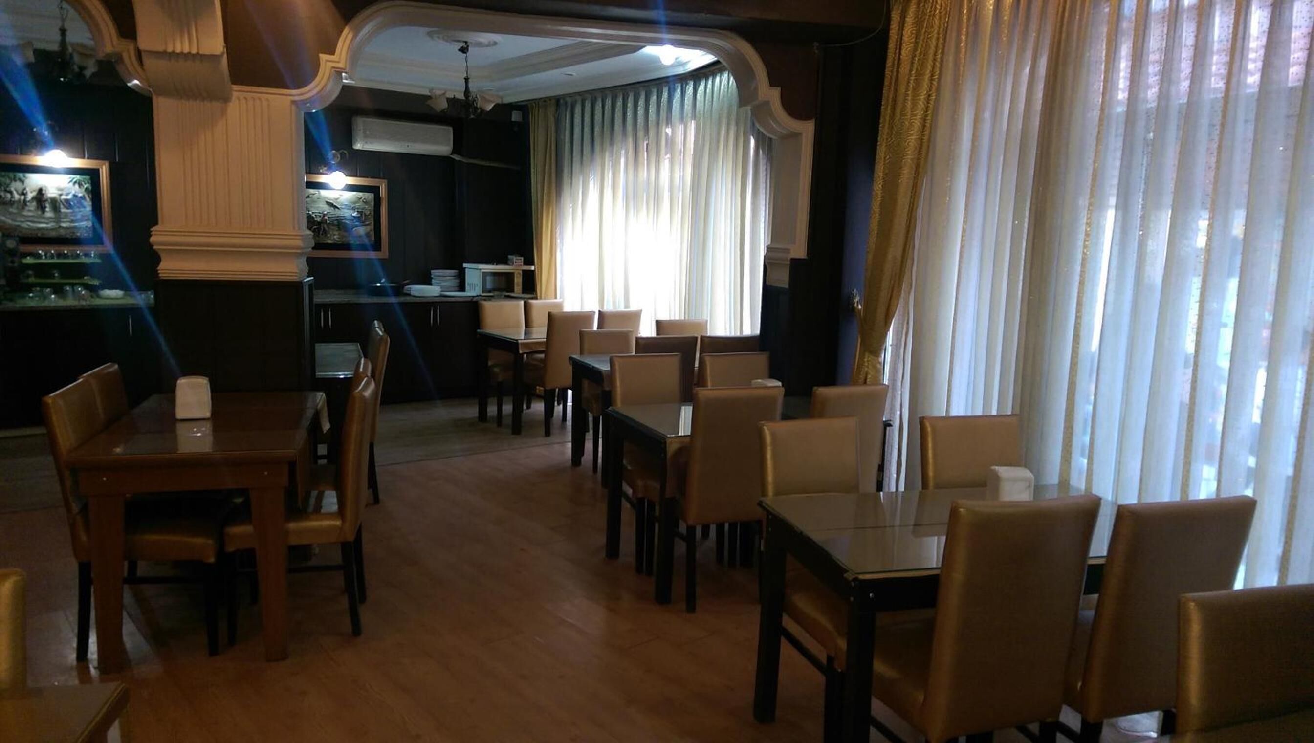 A dining area with wooden tables and chairs, a kitchen in the background, and a large window with curtains.