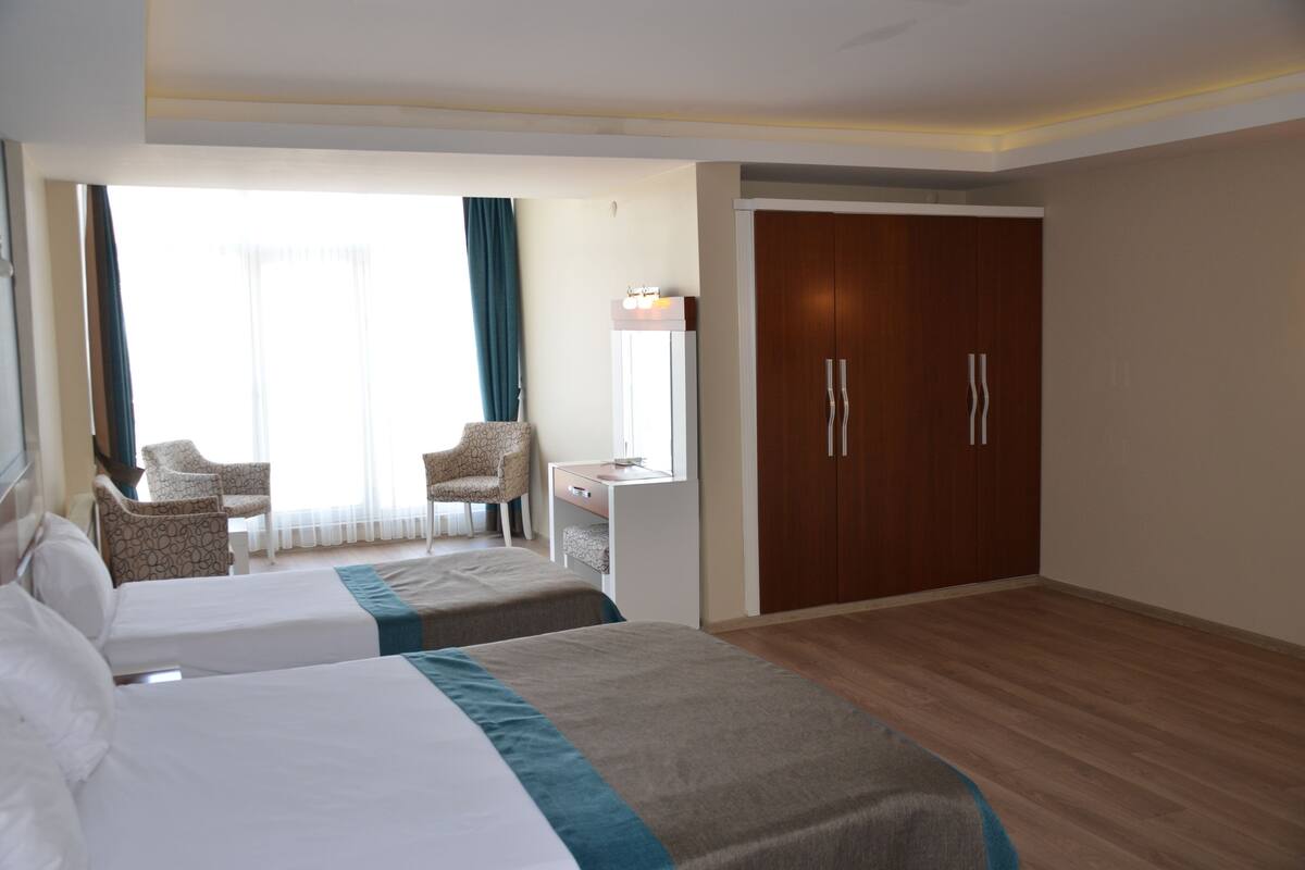Une chambre d’hôtel avec deux lits, un bureau, une chaise, une armoire et une grande fenêtre avec des rideaux.