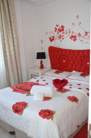A neatly made bed with a red rose pattern, a red headboard, and a small bedside table with a lamp.